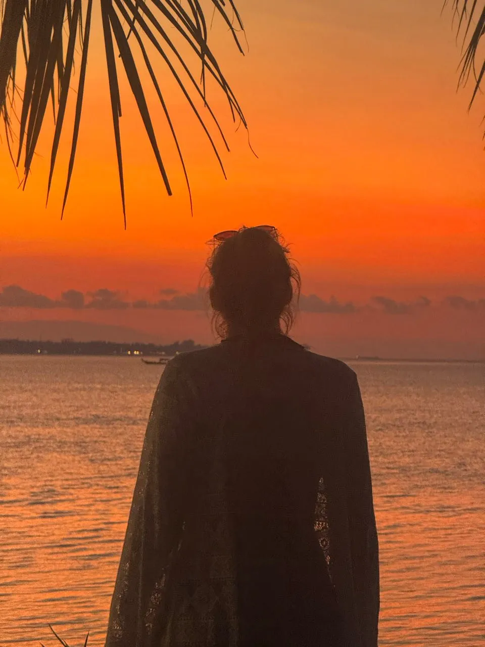 Silhouette einer Frau mit Haarknoten unter einem Palmwedel vor orangem Himmel, am fernen Ufer einzelne Lichter