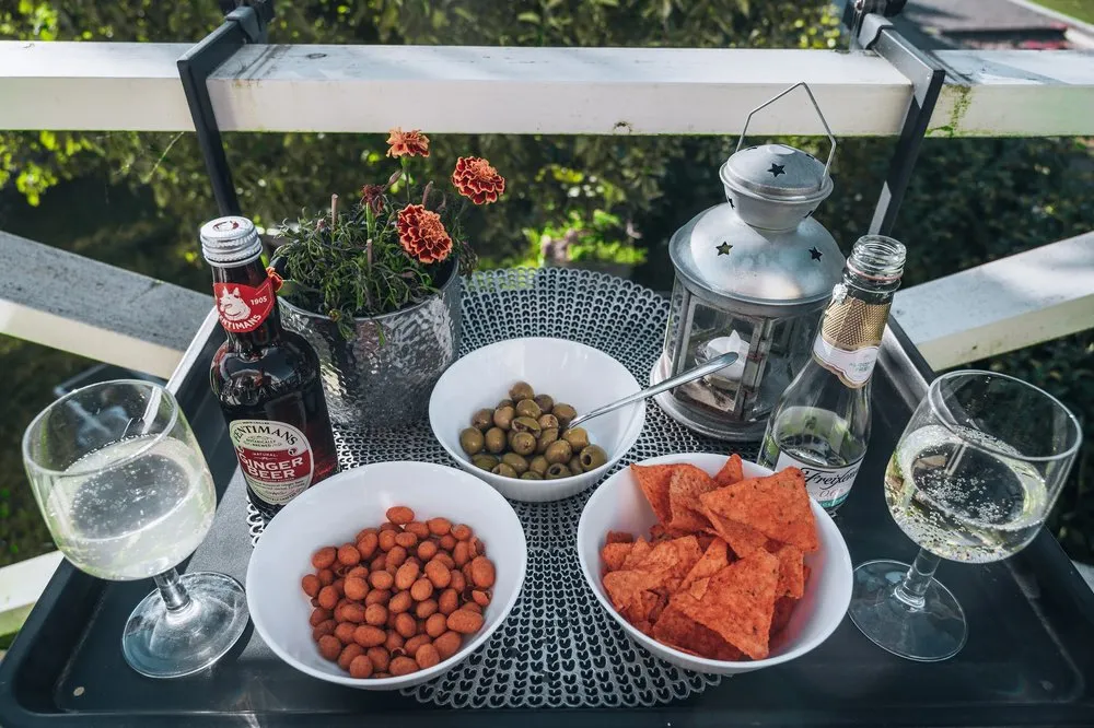 Tisch mit Ginger Beer, zwei Gläsern und Schalen mit Nüssen, Oliven und Chips, dahinter Balkongeländer und ein Blumentopf