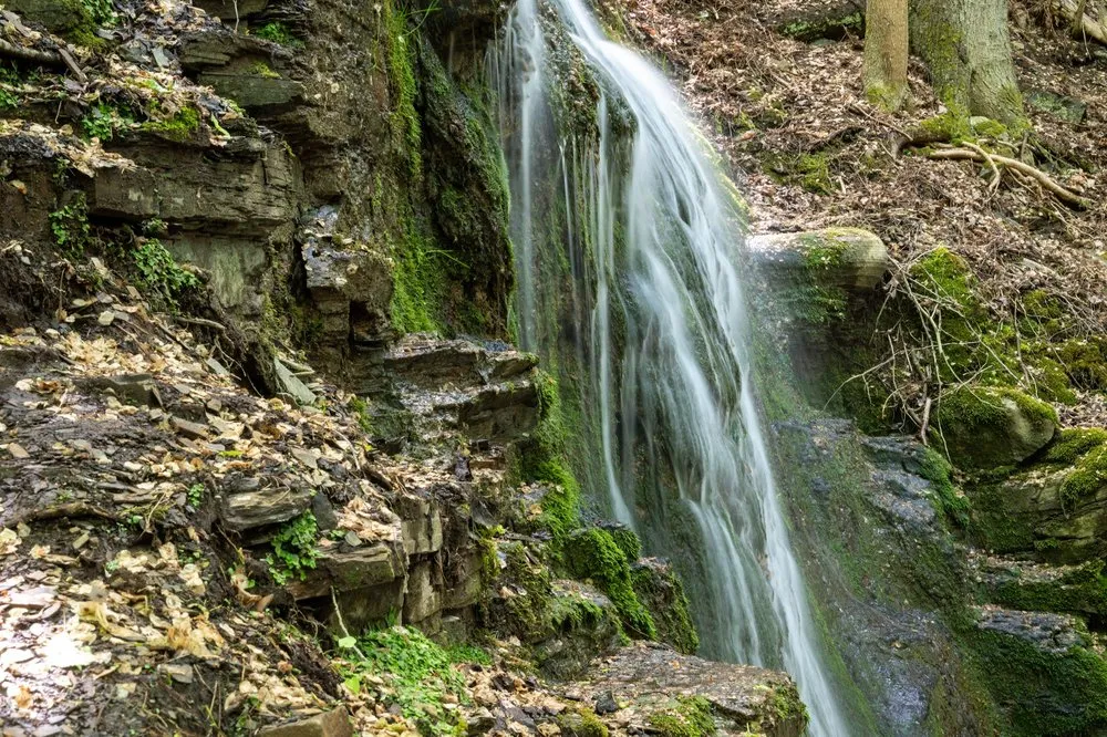 Wasserfall an einer moosigen Felswand, links ein Hang mit Laub und Steinplatten