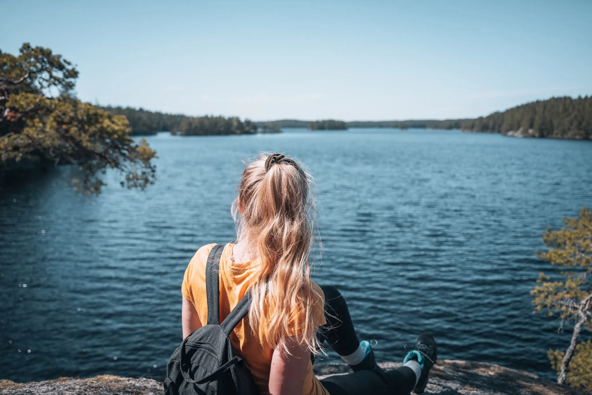 Frau mit Rucksack sitzt auf einem Felsen im Tiveden Nationalpark und blickt über einen See mit bewaldeten Ufern