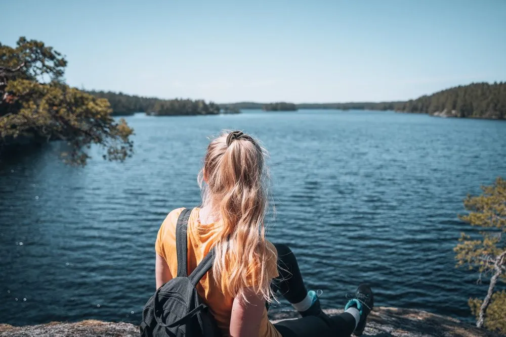 Frau mit Rucksack sitzt auf einem Felsen im Tiveden Nationalpark und blickt über einen See mit bewaldeten Ufern