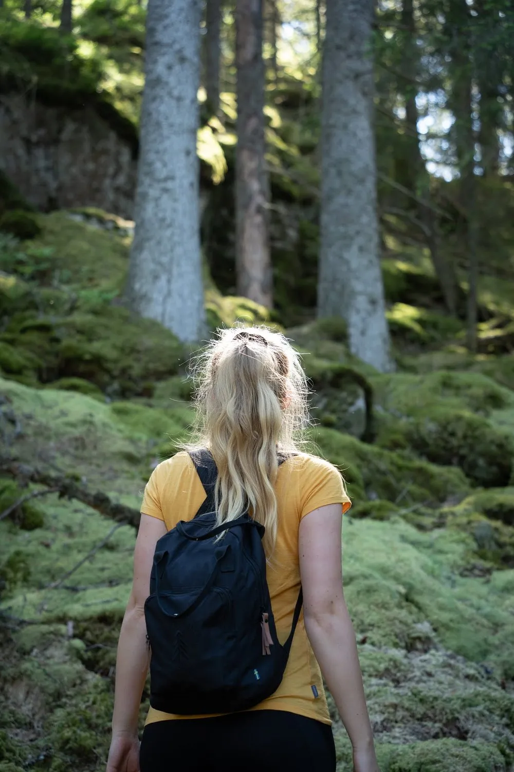 Frau mit gelbem Shirt und schwarzem Rucksack von hinten vor einem moosbewachsenen Hang mit Fichten