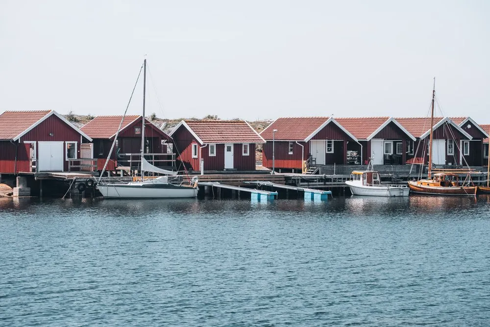 Dunkelrote Bootshäuser an einem Steg, davor Segelboote und ein Motorboot am glatten Wasser