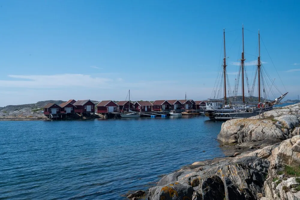 Rote Bootshäuser am Ufer einer Bucht, daneben liegt ein Schiff mit drei hohen Masten