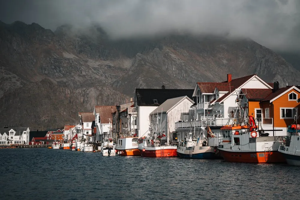 Fischerboote liegen im Hafen von Henningsvær vor den weissen und orangen Häusern, darüber dunkle Wolken an den Bergen