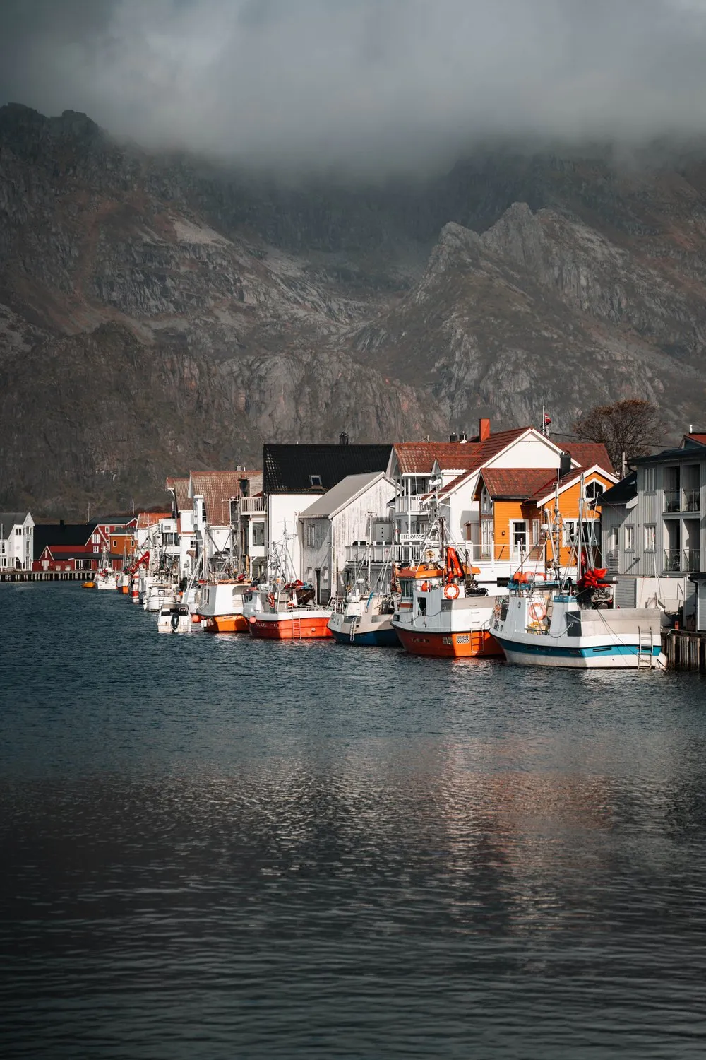 Eine lange Reihe Fischerboote vor den Häusern im Hafen von Henningsvær, darüber Wolken an den Bergen