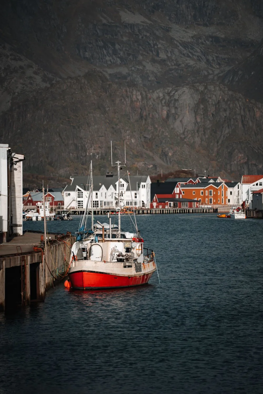 Ein rot-weisses Fischerboot an einer Holzmole in Henningsvær, gegenüber die weissen und orangen Häuser