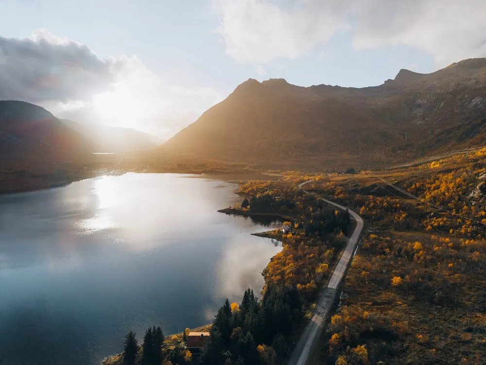 Eine Strasse führt am Ufer eines Fjords entlang durch gelbes Herbstlaub, dahinter die tiefe Sonne über den Bergen, aus der Luft