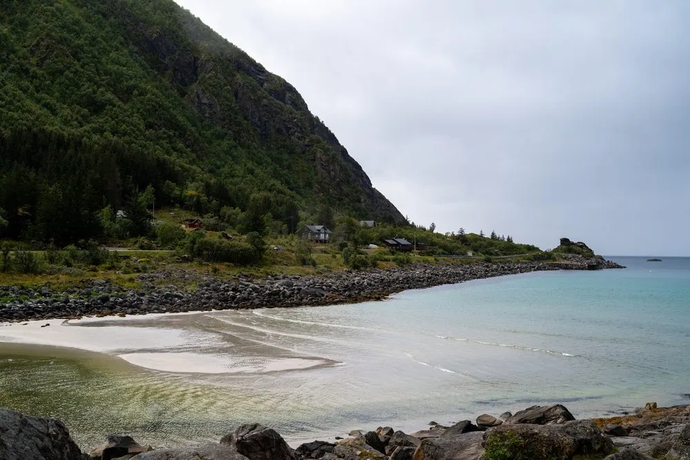Heller Sandstrand mit türkisfarbenem Wasser am Rørvikstranda, links ein bewaldeter Hang mit einzelnen Häusern