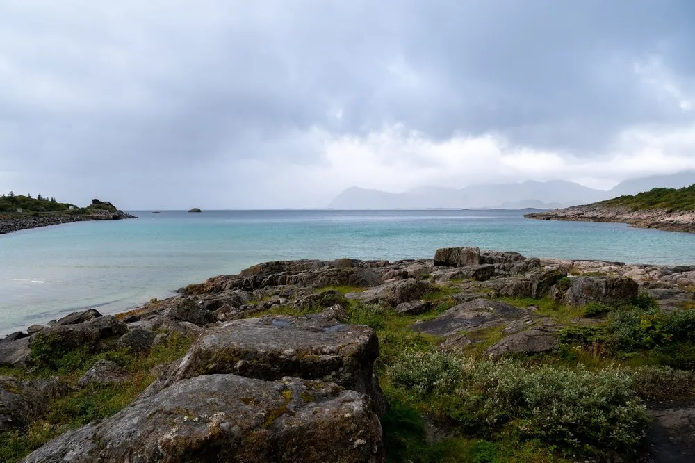 Felsiges Ufer und türkisfarbenes Wasser am Rørvikstranda unter dunklen Wolken, im Hintergrund Berge im Dunst