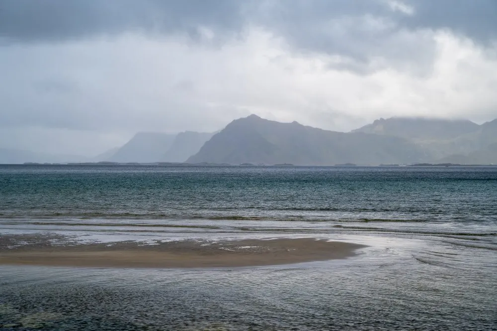 Flache Sandbank im Wasser, dahinter Berge im Regendunst