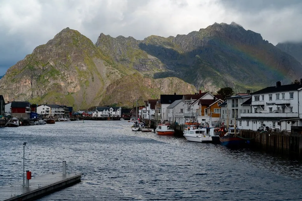 Der Hafen von Henningsvær mit Fischerbooten und weissen Häusern, dahinter ein Berg mit einem Regenbogen