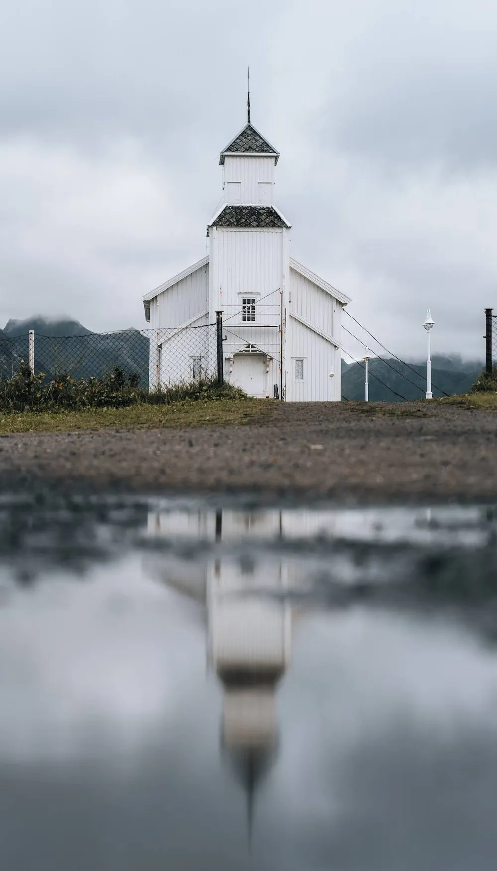 Die weisse Holzkirche von Gimsøy mit spitzem Turm, ihr Spiegelbild liegt in einer Pfütze im Vordergrund