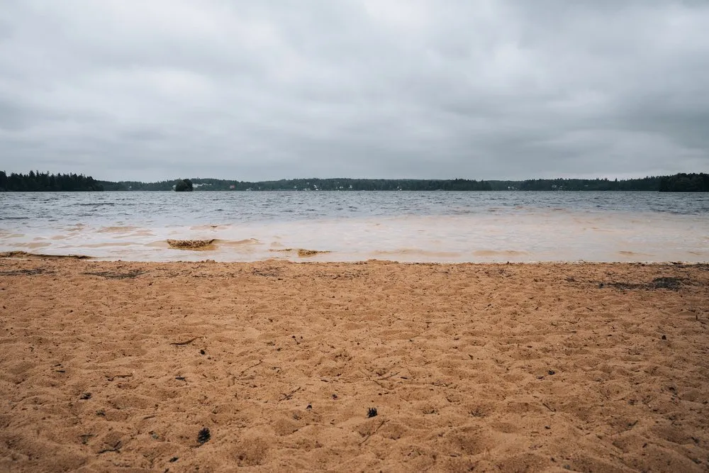 Sandstrand am See bei bedecktem Himmel, am Ufer färbt aufgewirbelter Grund das Wasser braun