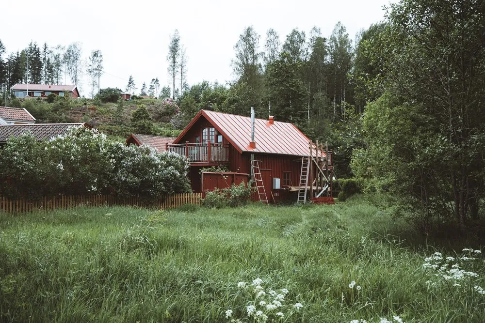 Ein rotes Holzhaus mit Balkon am Waldrand, davor eine hohe Wiese mit weissen Blumen