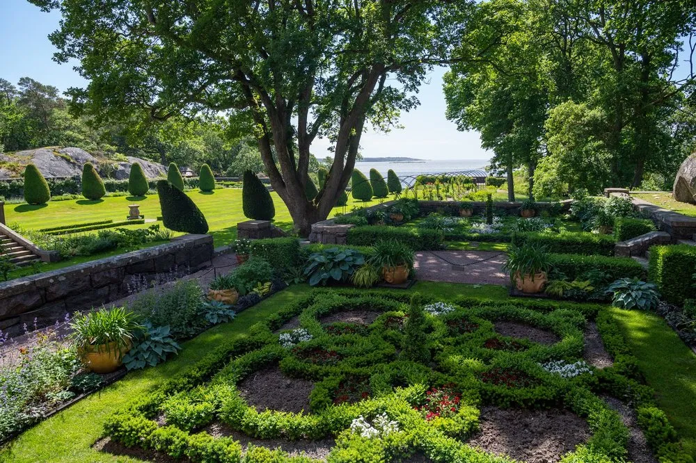 View from above of a geometric bed with clipped hedges, lawn terraces, large trees and the sea behind