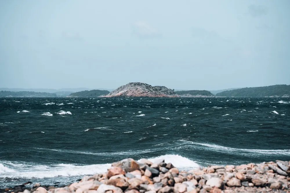 Dark, churning sea with white crests in front of a rocky island, stones in the foreground