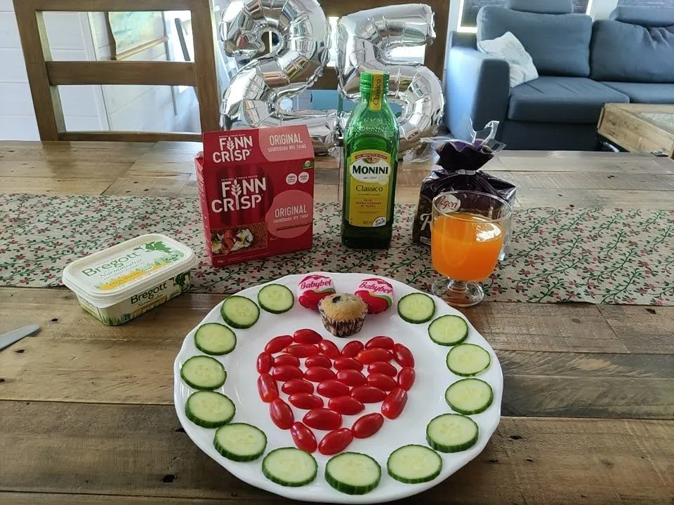 Frühstückstisch mit Tomatenherz und Gurkenscheiben auf dem Teller, Muffin, Knäckebrotpackung, Butter, Orangensaft und silbernen Ballons in Form einer 25