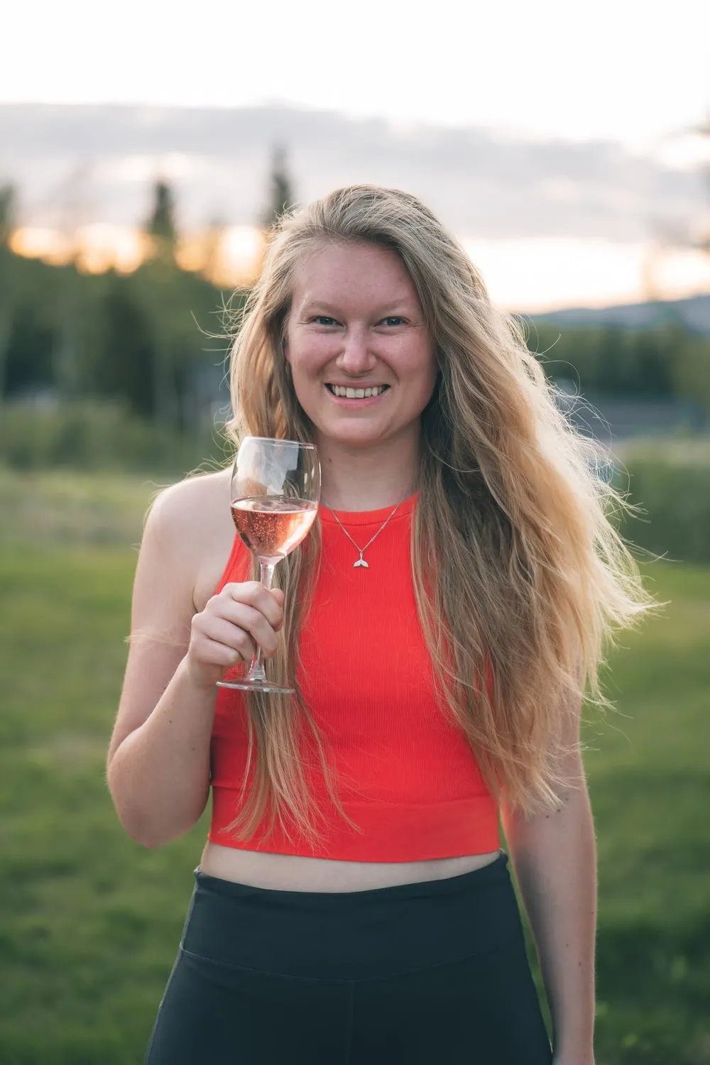 Annina in rotem Top hält lachend ein Glas Rosé, hinter ihr Wiese und Wald im Abendlicht
