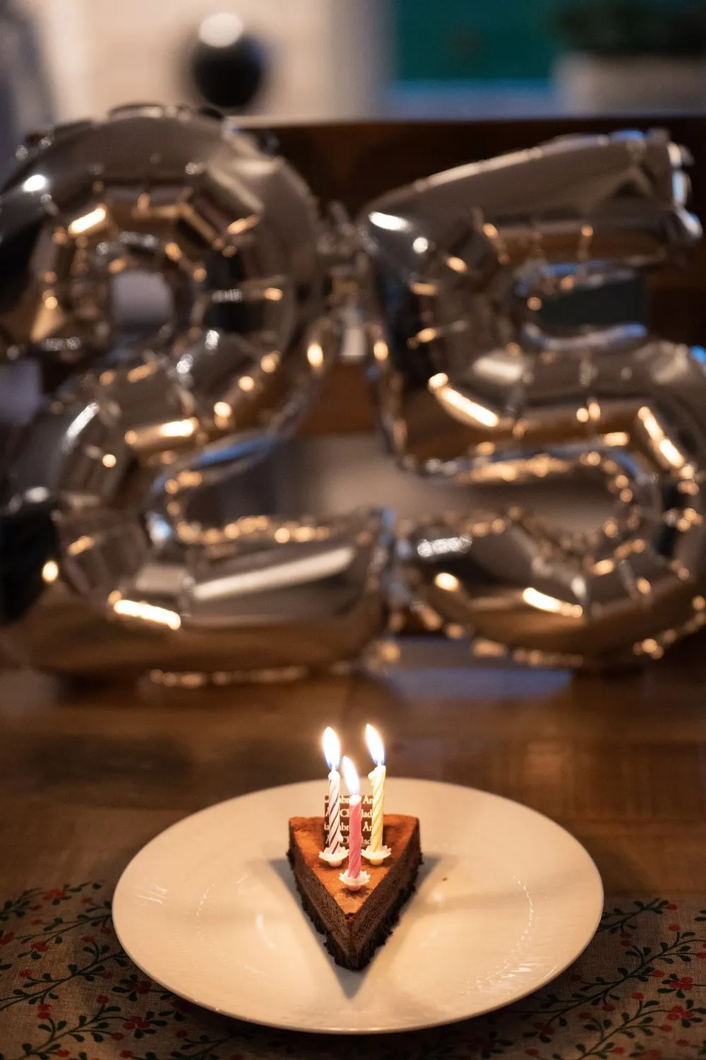 Ein Stück Schokoladenkuchen mit drei brennenden Kerzen vor silbernen Ballons in Form einer 25