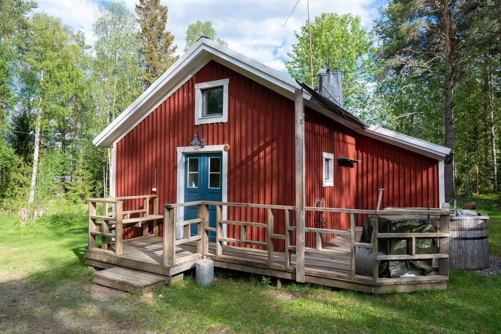 Rotes Holzhaus mit blauer Tür und Holzveranda, umgeben von Birken und Föhren