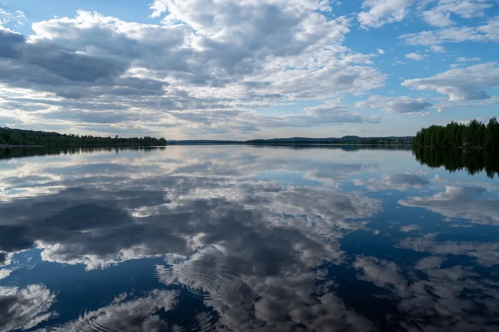 Wolken und blauer Himmel spiegeln sich im glatten Wasser des Sees bei Rätan