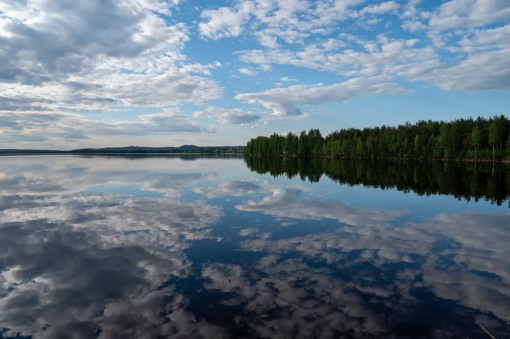 Ruhiger See mit Waldufer rechts, das sich samt den Wolken im Wasser spiegelt