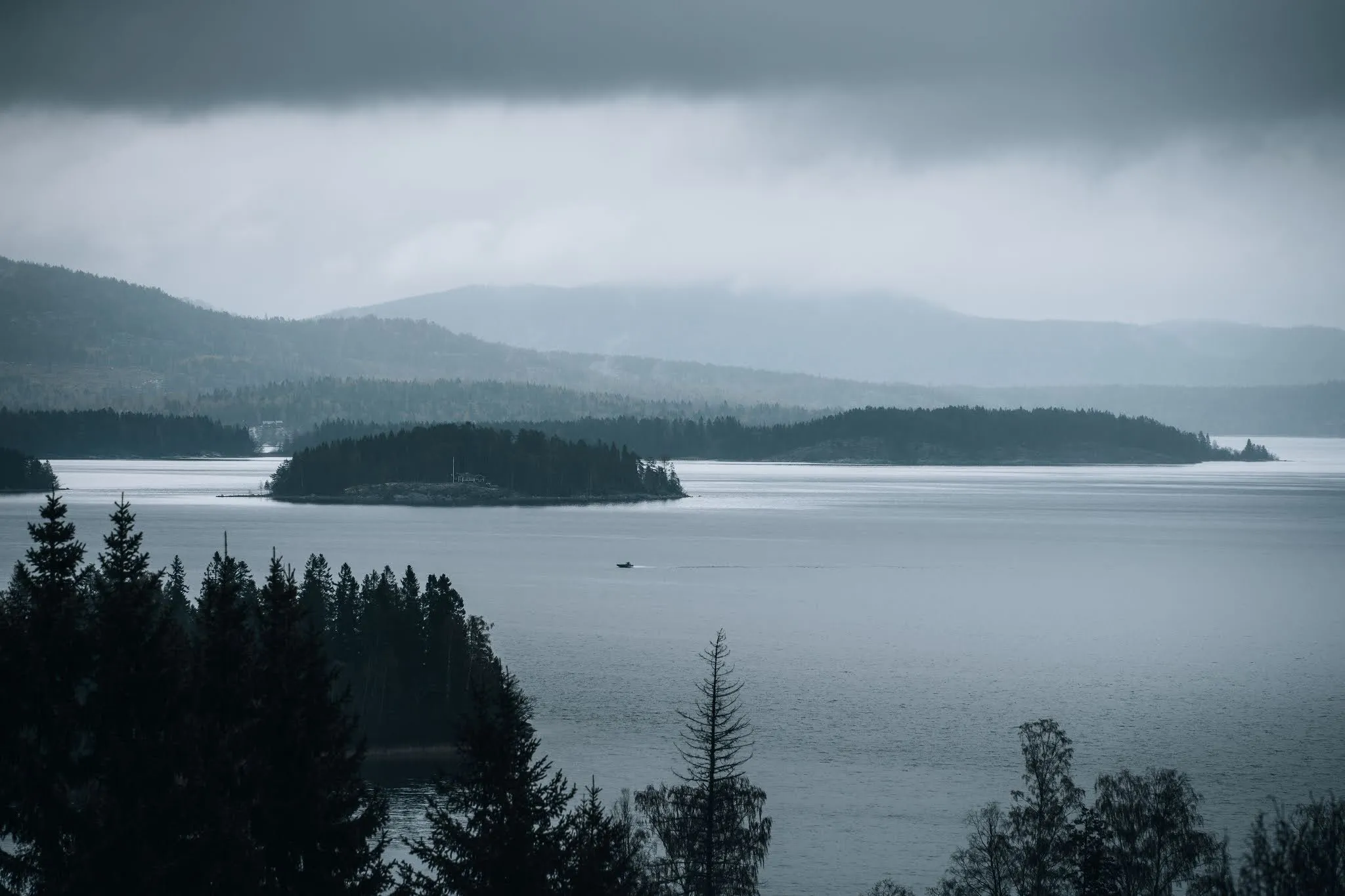 Bewaldete Insel in einer grauen Wasserfläche unter tiefen Regenwolken, dahinter Hügel im Dunst