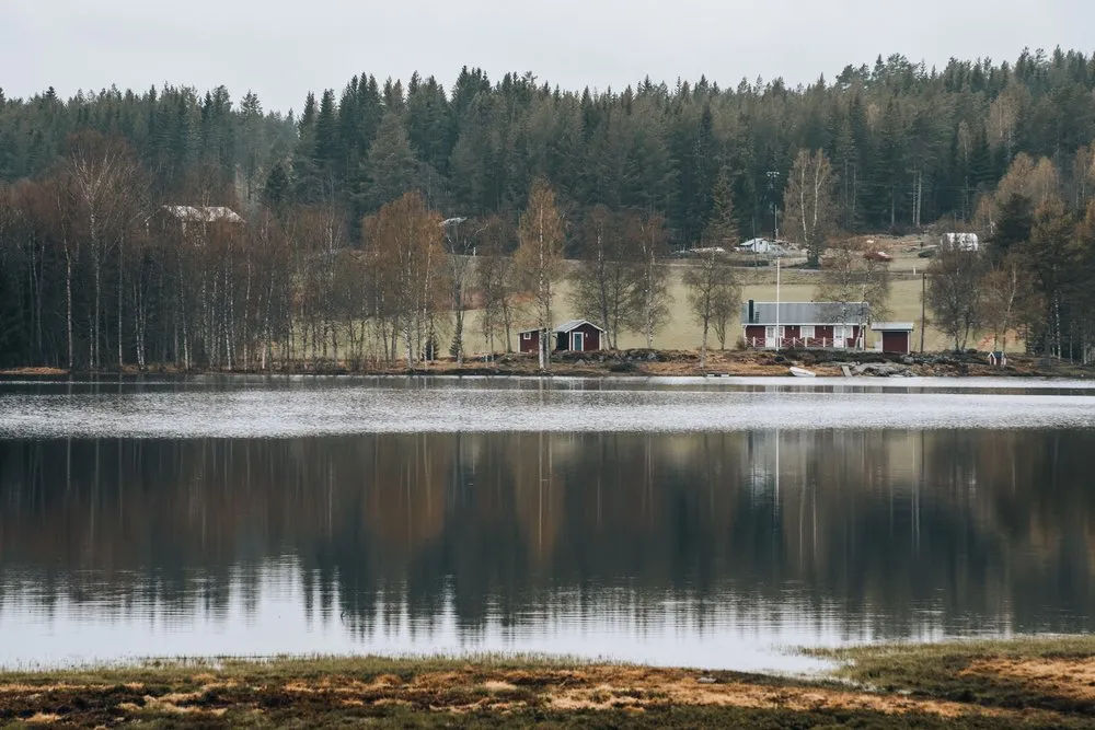 Rote Holzhäuser am Ufer eines Sees vor dunklem Nadelwald, die kahlen Birken spiegeln sich im Wasser