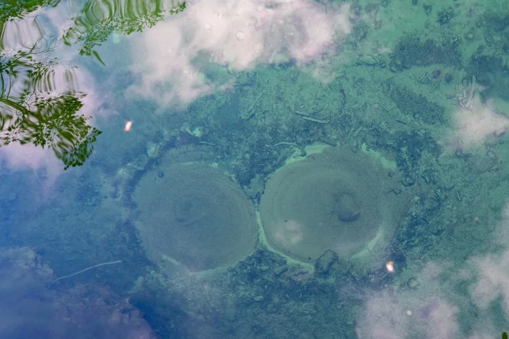 Zwei runde Quellöffnungen auf dem Grund des Sees, darüber spiegeln sich Wolken