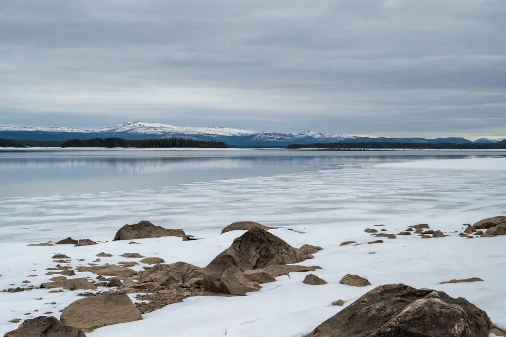 Grosse Felsbrocken am Ufer eines halb zugefrorenen Sees, am Horizont schneebedeckte Berge