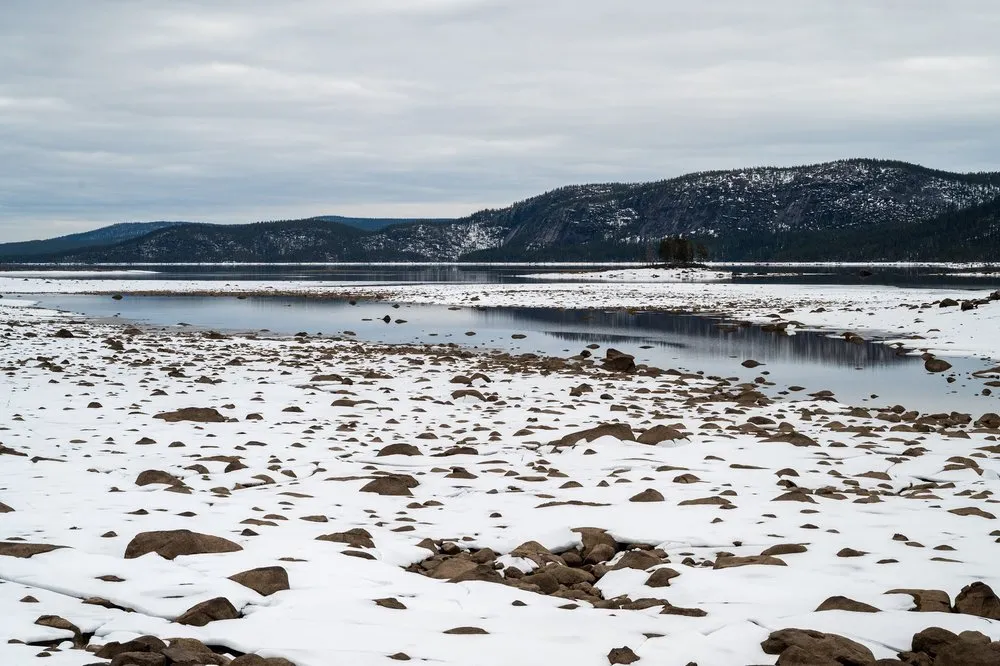 Flache Uferzone mit Schnee und Steinen, dahinter ein Wasserarm und ein bewaldeter Bergrücken