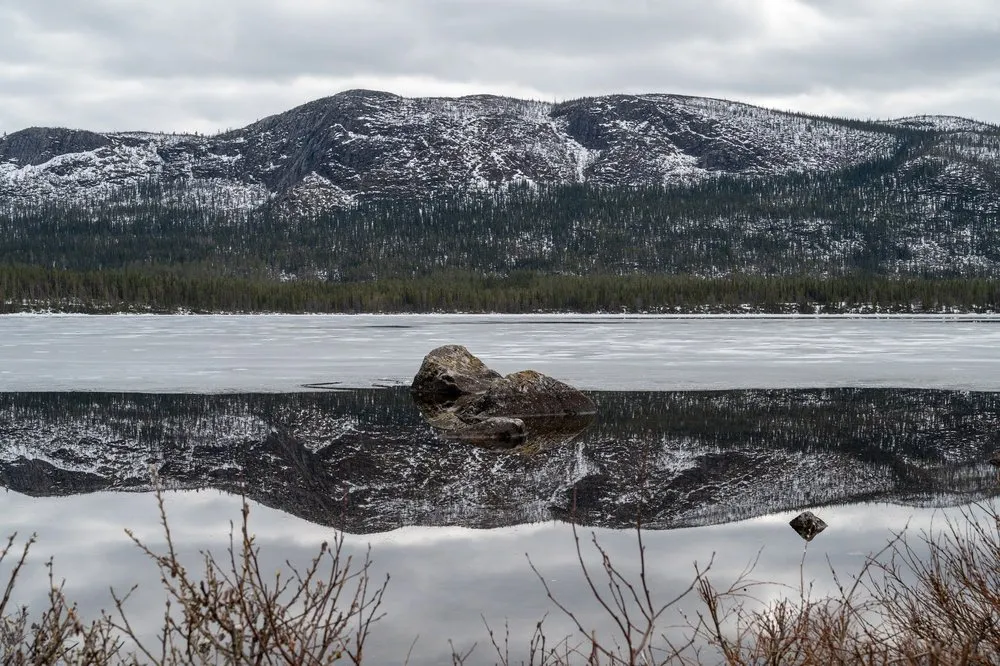 Ein Felsblock ragt aus dem spiegelglatten Wasser, darin das Spiegelbild eines schneebedeckten Berghangs