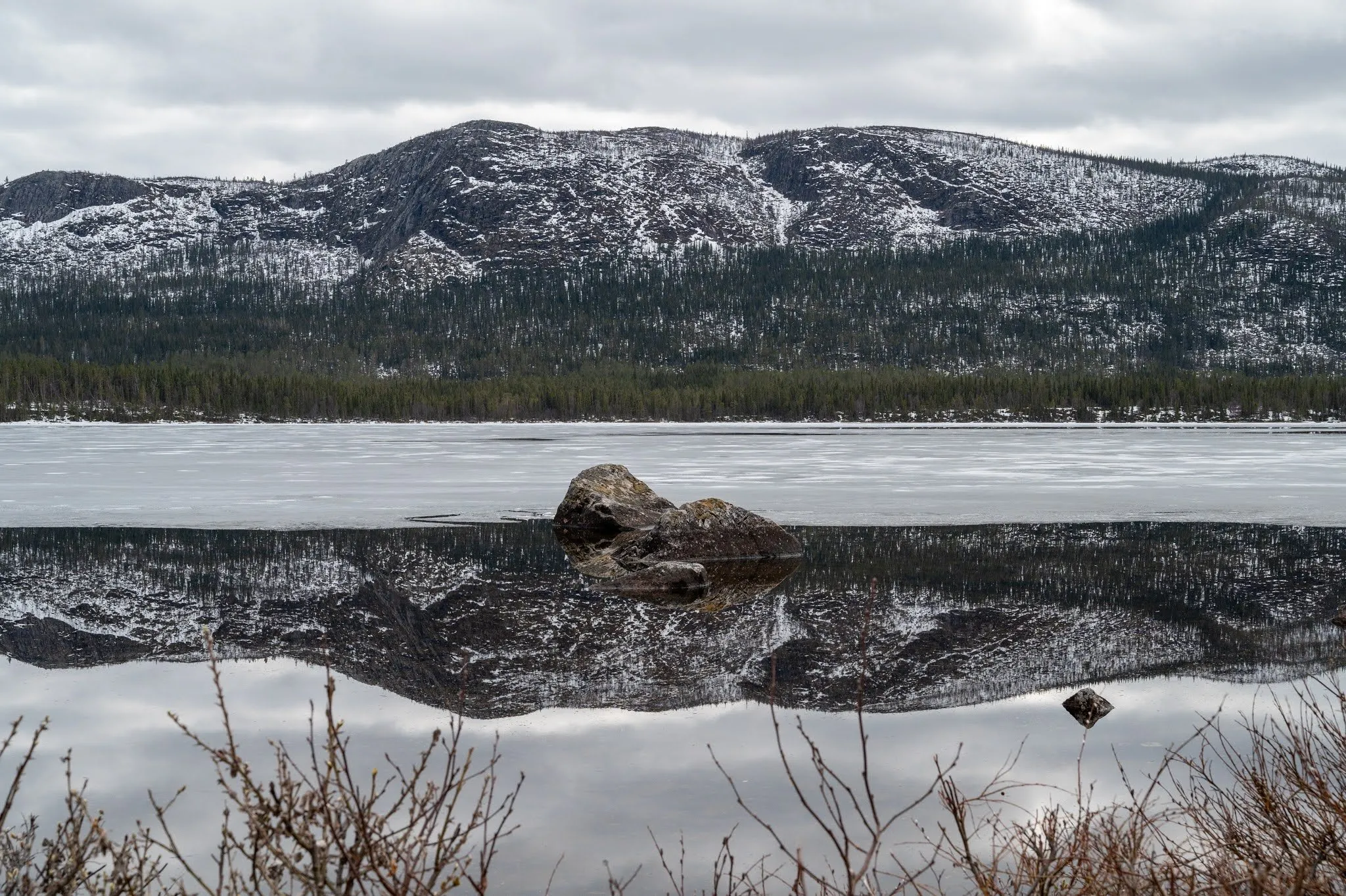 Ein Felsblock ragt aus dem spiegelglatten Wasser, darin das Spiegelbild eines schneebedeckten Berghangs