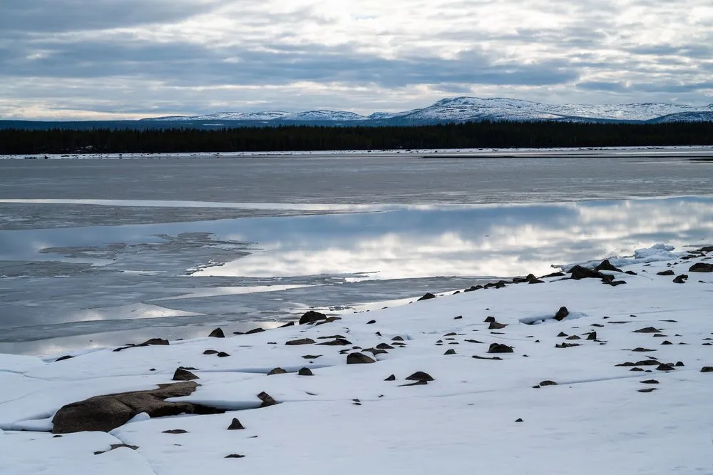 Verschneites Ufer mit dunklen Steinen, dahinter eine spiegelnde Wasserfläche und schneebedeckte Berge am Horizont