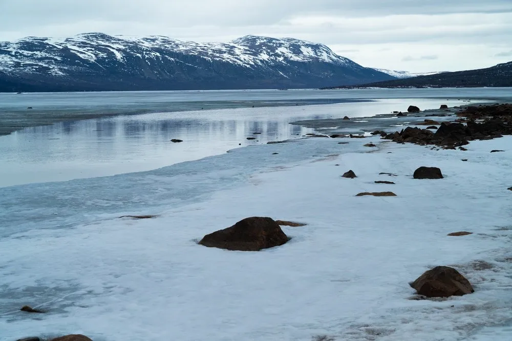 Steine ragen aus Schnee und Eis am Ufer, dahinter offenes Wasser und ein Berg mit Schnee
