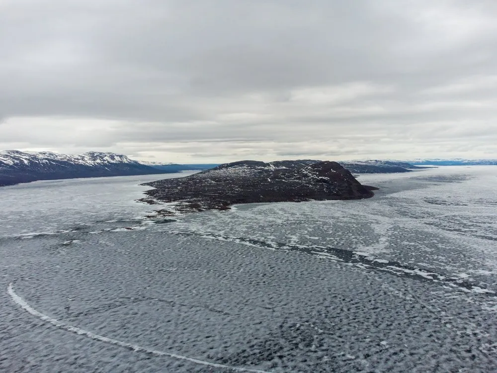 Drohnenblick auf eine dunkle Landzunge im zugefrorenen See, links Berge mit Schneeresten