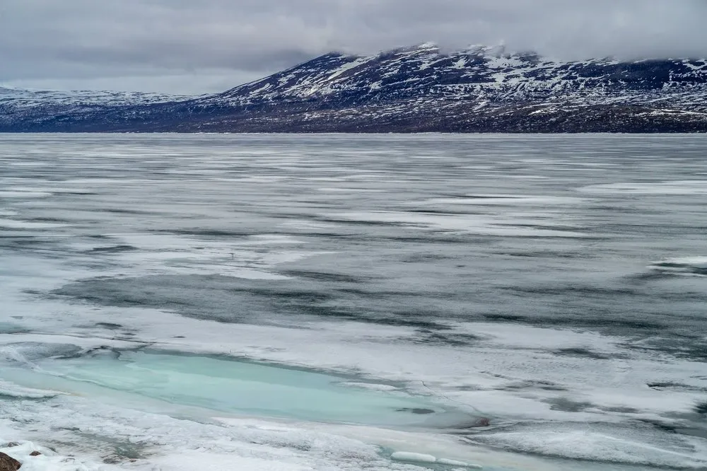 Eisfläche mit einer türkisfarbenen Stelle im Vordergrund, dahinter ein Berg mit Schneefeldern unter tiefen Wolken