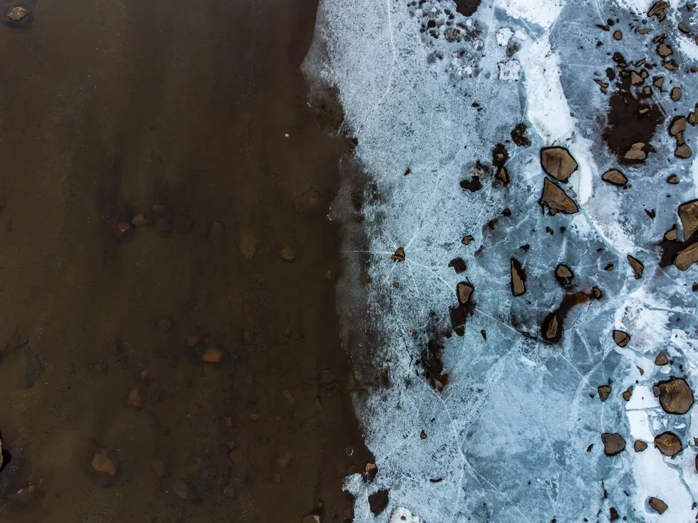 Drohnenfoto senkrecht nach unten: links dunkles, eisfreies Wasser, rechts rissiges Eis mit dunklen Steinen