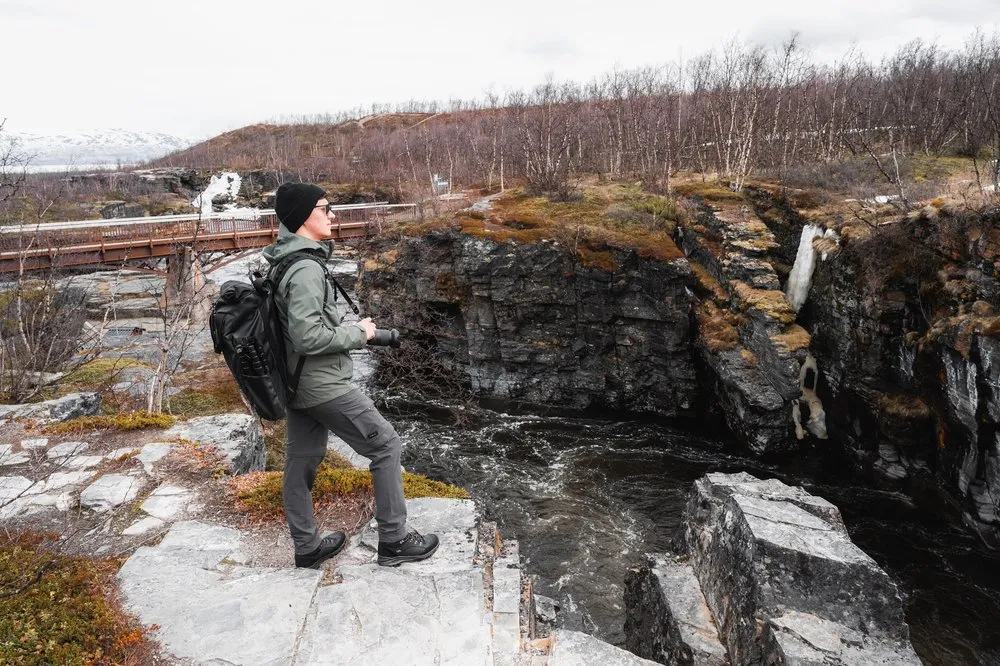 Eine Person mit Rucksack und Kamera steht am Wasserfall im Abisko Nationalpark auf Felsplatten über der Schlucht, hinten eine Brücke