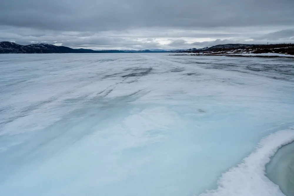 Weite, hellblau schimmernde Eisfläche eines Sees, am Horizont dunkle Hügel unter grauem Himmel