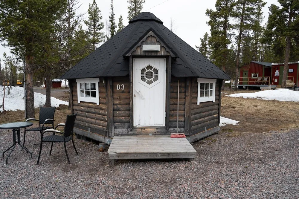 Unser Häuschen auf dem Campingplatz in der Nähe von Kiruna: ein kleines Holzhaus mit dunklem Dach und weisser Tür, davor Tisch und Stühle auf Kies