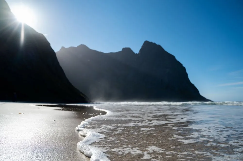 Gegenlicht über einer dunklen Felswand, im Vordergrund läuft eine Welle mit Schaum über nassen Sand
