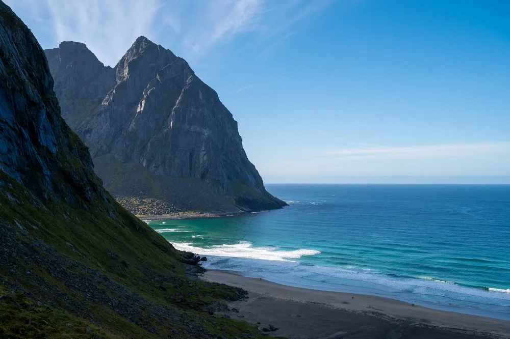 Blick auf die Kvalvika Beach: eine steile Felswand über türkisfarbenem Meer und Sandstrand bei blauem Himmel