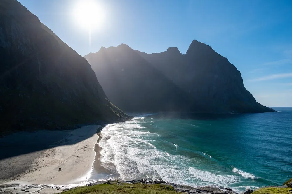 Sandstrand mit Brandung und einzelnen Personen unterhalb der Felswände, darüber die Sonne