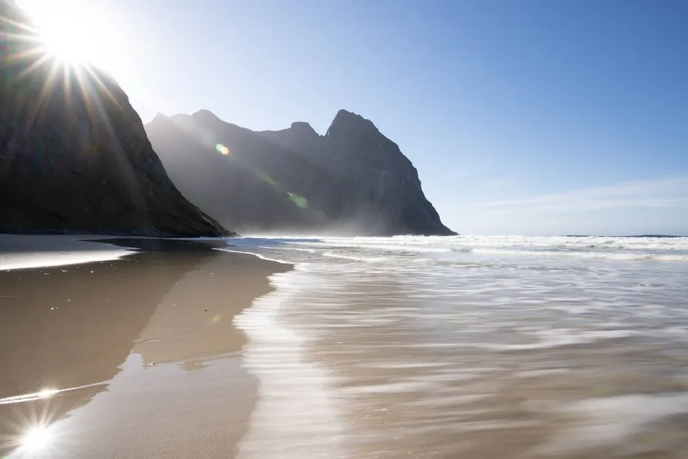 Auslaufende Wellen in Langzeitbelichtung auf nassem Sand, die Sonne strahlt hinter dem Fels