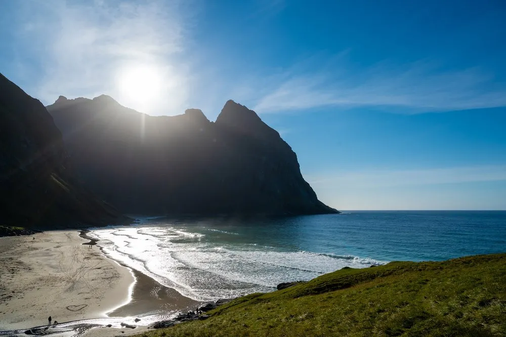 Blick über eine grüne Hangkante auf den Strand, die Sonne steht direkt über dem Bergkamm