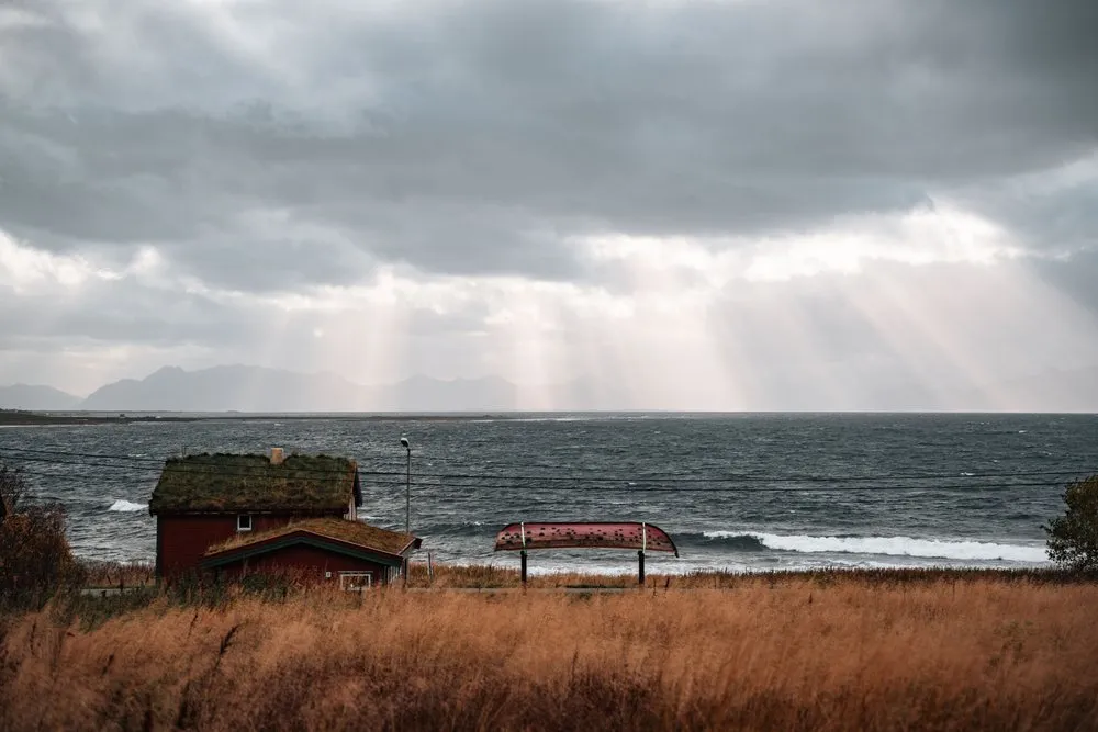 Sonnenstrahlen brechen durch dunkle Sturmwolken über dem aufgewühlten Meer bei Nøss, vorne ein Haus mit Grasdach und trockenes Gras