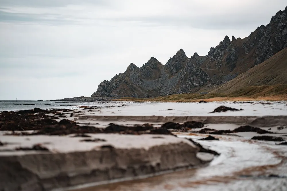 Der helle Sandstrand von Bleik mit dunklem Tang und dem Bach, der sich durch den Sand zieht, dahinter der gezackte Grat