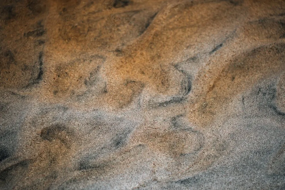 Rostbraune und dunkle, geschwungene Muster im Wasser des eisenhaltigen Bachs am Strand von Bleik